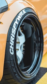 Close-up of a car tire featuring white lettering and a visible brake caliper. The wheel has a matte black finish with a glossy lip and the vehicle has a bright orange paint job.