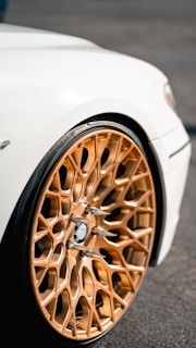 A close-up view of a car's intricate and stylish golden alloy wheel against a white vehicle body. The wheel features a complex geometric pattern with metallic elements and spike-like accents, all of which highlight the luxury sports car's design.