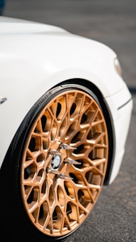 A close-up view of a car's intricate and stylish golden alloy wheel against a white vehicle body. The wheel features a complex geometric pattern with metallic elements and spike-like accents, all of which highlight the luxury sports car's design.