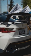 A sleek white sports car is shown from the rear, featuring a prominent spoiler and custom rear lights. Two flags with text are mounted on the car. The license plate is visible with an Oregon registration. The background includes blurred greenery and utility poles, indicating an outdoor setting.