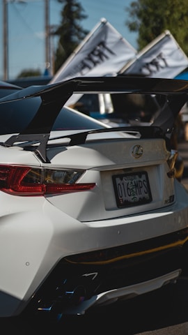 A sleek white sports car is shown from the rear, featuring a prominent spoiler and custom rear lights. Two flags with text are mounted on the car. The license plate is visible with an Oregon registration. The background includes blurred greenery and utility poles, indicating an outdoor setting.