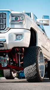 A close-up view of the front section of a lifted white pickup truck with large, rugged tires and a shiny chrome grille. The suspension system features visible red components, and the background shows a clear blue sky.