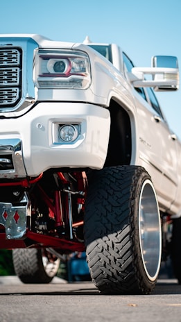 A close-up view of the front section of a lifted white pickup truck with large, rugged tires and a shiny chrome grille. The suspension system features visible red components, and the background shows a clear blue sky.