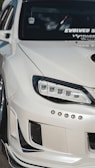 A close-up view of the front left portion of a modified white car, showcasing custom headlights and bumper details. The windshield features multiple decals, and the car's exterior has a glossy finish.