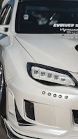 A close-up view of the front left portion of a modified white car, showcasing custom headlights and bumper details. The windshield features multiple decals, and the car's exterior has a glossy finish.