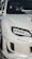 A close-up view of the front left portion of a modified white car, showcasing custom headlights and bumper details. The windshield features multiple decals, and the car's exterior has a glossy finish.