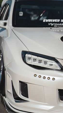 A close-up view of the front left portion of a modified white car, showcasing custom headlights and bumper details. The windshield features multiple decals, and the car's exterior has a glossy finish.