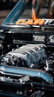 A close-up of a car engine with a detailed view of the metal components and pipes. The hood is open, and the engine is prominently displayed, showcasing its metallic and industrial characteristics.