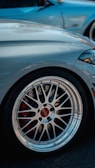 Close-up of a car's wheel with a shiny, detailed alloy rim. The brake system is slightly visible through the spokes, with a red caliper adding contrast. The car is a sleek, silvery color with another blue vehicle partially visible in the background.