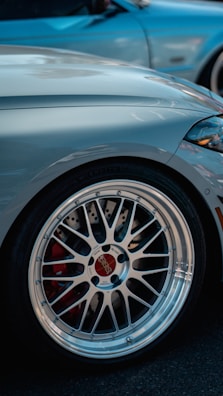 Close-up of a car's wheel with a shiny, detailed alloy rim. The brake system is slightly visible through the spokes, with a red caliper adding contrast. The car is a sleek, silvery color with another blue vehicle partially visible in the background.