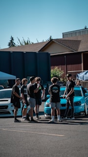 A group of young people are gathered outdoors on a sunny day. They are standing on a paved area near two parked cars, one of which is a bright blue, modified sports car. The background features residential-style buildings and some trees.