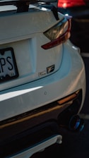A close-up view of the rear end of a white sports car featuring a prominent rear spoiler and dual exhaust. The car's badge displays 'RC F,' indicating the model. Shadows and reflections play across the car's surface, suggesting it is parked in a shaded or partially sunlit area. Another vehicle, partially visible, appears red in color in the background.
