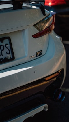 A close-up view of the rear end of a white sports car featuring a prominent rear spoiler and dual exhaust. The car's badge displays 'RC F,' indicating the model. Shadows and reflections play across the car's surface, suggesting it is parked in a shaded or partially sunlit area. Another vehicle, partially visible, appears red in color in the background.