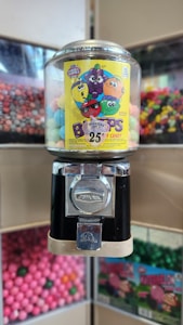 A vintage-style gumball machine with a transparent plastic globe filled with colorful candies. A bright yellow sticker with cartoon fruit characters is displayed on the front, advertising 25-cent treats. The background features other candy dispensers filled with different colored treats.