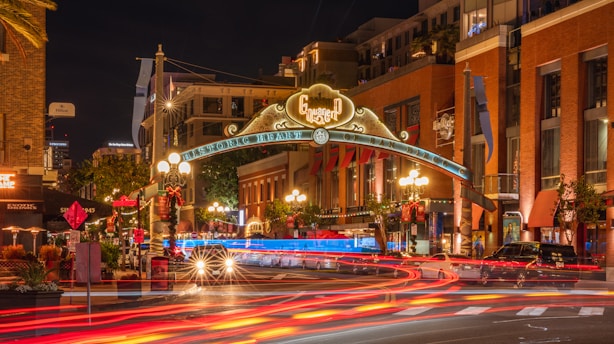 An illuminated urban street scene at night features a prominent archway with decorative lighting that welcomes visitors to the historic district. The street is lined with vibrant buildings and bustling traffic, creating trails of light from moving vehicles. Brightly lit signage and ambient street lights add to the lively atmosphere.