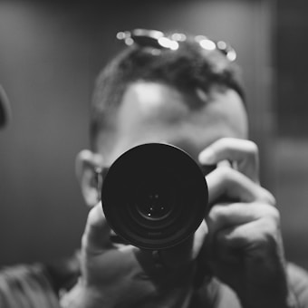 A person is holding a camera close to their face, with the lens pointed directly at the viewer. The composition is in black and white, giving it a classic and timeless feel. The person's head is partially visible, with short hair and glasses resting on top. The focus is on the camera lens, conveying a sense of intimacy and curiosity.
