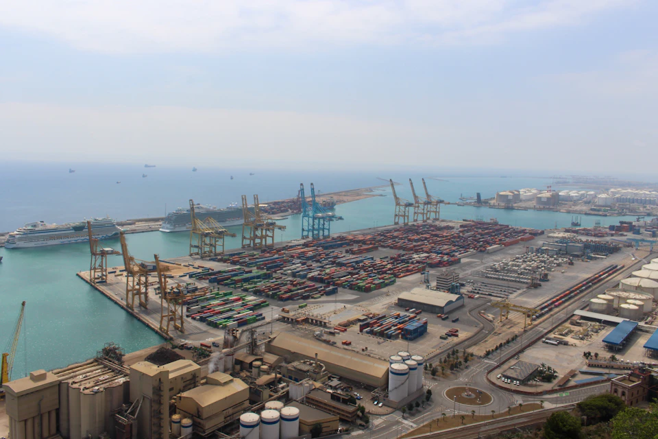 The image presents an expansive view of a bustling port city, teeming with activity and filled with numerous cranes and ships. The city is nestled on the coast, with the vast expanse of the ocean stretching out to meet it at the horizon. The sky above is a clear blue, dotted with fluffy white clouds that add depth to the scene.

The port itself is a hive of activity, with multiple cranes standing tall and ready for use, their long arms reaching towards the sky as they load and unload cargo from ships. These cranes are painted in shades of green and yellow, adding a vibrant touch to the industrial landscape. The cityscape is dotted with buildings of various sizes, some taller than others, all contributing to the overall grandeur of the scene.

The image captures the essence of urban life at sea level, showcasing the intricate network of roads that crisscrosses the city and connects it to other ports around the world.