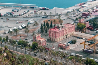 A coastal area with a large, pink historical building surrounded by palm trees and parking spaces. The scene includes several trucks and containers arranged in a nearby shipping area. The blue ocean stretches into the background, and the overall setup suggests an industrial or commercial port environment.