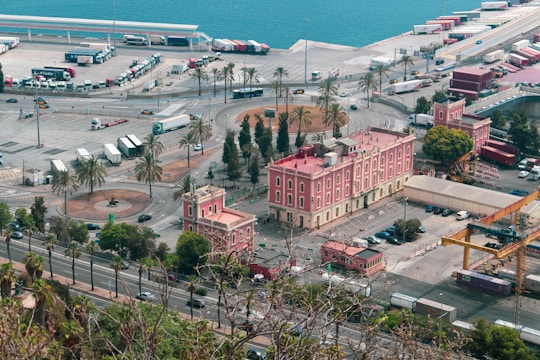 A coastal area with a large, pink historical building surrounded by palm trees and parking spaces. The scene includes several trucks and containers arranged in a nearby shipping area. The blue ocean stretches into the background, and the overall setup suggests an industrial or commercial port environment.