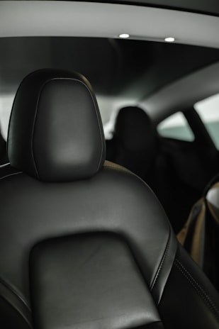 A sleek, modern car interior featuring a black leather seat with visible stitching. The minimalist design includes a smooth black seatback and headrest. Overhead, ambient lighting is installed in the ceiling, providing a subtle, elegant illumination. The background is slightly out of focus, suggesting a compact and cozy space.