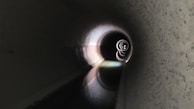 A long, dark tunnel with a rounded shape, featuring concentric rings of light that gradually become smaller towards the center. The tunnel has textured walls and a reflective surface on the floor, creating an intriguing play of light and shadow.