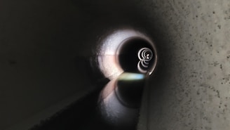 A long, dark tunnel with a rounded shape, featuring concentric rings of light that gradually become smaller towards the center. The tunnel has textured walls and a reflective surface on the floor, creating an intriguing play of light and shadow.