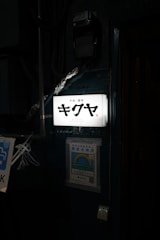 An illuminated sign with Japanese characters and the word 'Kikuya' and 'Shinjuku' below it. The sign is attached to a wall which has a poster with a rainbow and Japanese text below the sign. Various pipes and wires are visible around the sign.
