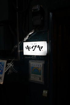 An illuminated sign with Japanese characters and the word 'Kikuya' and 'Shinjuku' below it. The sign is attached to a wall which has a poster with a rainbow and Japanese text below the sign. Various pipes and wires are visible around the sign.