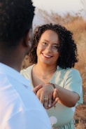 A woman with curly hair smiles warmly while holding hands with another person. She is wearing a light green dress with white polka dots and a brown bracelet. The background is a blurred natural setting with dried grass and soft lighting.