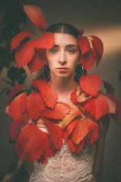 A woman with dark hair, wearing large hoop earrings and a lace dress, gazes directly at the camera. Her head and upper body are adorned with vibrant red leaves, creating a striking contrast with her outfit.