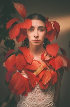 A woman with dark hair, wearing large hoop earrings and a lace dress, gazes directly at the camera. Her head and upper body are adorned with vibrant red leaves, creating a striking contrast with her outfit.