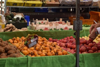 A market stall displays a variety of fresh fruits, including persimmons, peaches, avocados, and kiwis. The fruits are arranged on a green cloth, and a hand is seen selecting one of the fruits. Wooden crates and boxes with labels like 'Fresón de Palos' are visible in the background. Pineapples are in yellow boxes on a shelf above.