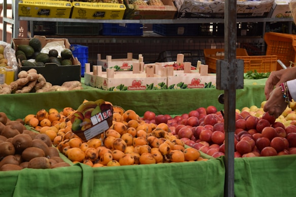 A market stall displays a variety of fresh fruits, including persimmons, peaches, avocados, and kiwis. The fruits are arranged on a green cloth, and a hand is seen selecting one of the fruits. Wooden crates and boxes with labels like 'Fres&oacute;n de Palos' are visible in the background. Pineapples are in yellow boxes on a shelf above.