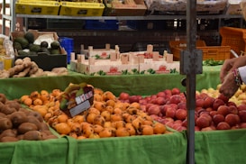 A market stall displays a variety of fresh fruits, including persimmons, peaches, avocados, and kiwis. The fruits are arranged on a green cloth, and a hand is seen selecting one of the fruits. Wooden crates and boxes with labels like 'Fresón de Palos' are visible in the background. Pineapples are in yellow boxes on a shelf above.