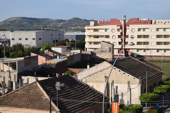 A suburban area with a mix of residential and industrial buildings. In the foreground, there are weathered, older houses with tiled roofs and peeling plaster. Behind them, a larger, modern apartment building with a red and beige facade stands out. In the background, an industrial facility with a large warehouse is visible. The scene is bordered by green trees and hills under a clear blue sky.