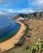 A coastal landscape featuring a sandy beach with a row of palm trees running along a road. Clear blue and green waters meet the shore, with a breakwater extending into the sea. A hillside with lush green vegetation rises steeply on the right side, and a town with white buildings is nestled in the valley near the coastline. In the distance, there are more mountains and a ship sailing near the horizon under a partly cloudy sky.