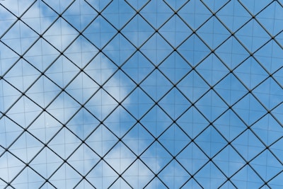 A geometric pattern composed of a series of triangular glass panes forming a large grid, with blue sky and scattered white clouds visible through the panels.