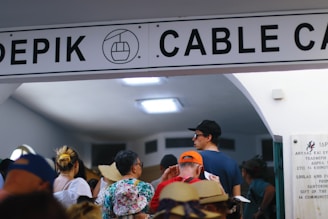 A group of people are waiting in line under a sign for a cable car station. The diverse crowd includes individuals wearing hats and casual clothing. The setting appears to be indoors with a white ceiling and fluorescent lights.