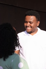 A man and a woman are interacting, with the man smiling warmly at the woman. The man is wearing a white polo shirt and has short, curly hair. The woman has curly hair and is wearing a green outfit with large white polka dots. The background is dark, contrasting with the light on their faces.