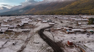 A rural settlement is covered with a light dusting of snow. The scene includes numerous small wooden buildings, dirt roads, and a few scattered vehicles. A backdrop of forested and mountainous terrain is visible, with clouds partially covering the sky.