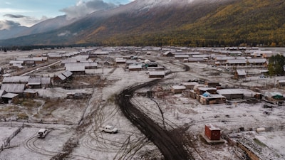 A rural settlement is covered with a light dusting of snow. The scene includes numerous small wooden buildings, dirt roads, and a few scattered vehicles. A backdrop of forested and mountainous terrain is visible, with clouds partially covering the sky.