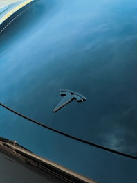A shiny, reflective car hood displaying a Tesla logo against a smooth, blue, and cloudy sky backdrop.