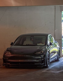 A sleek, dark-colored Tesla car is parked in a dimly lit concrete garage. The electric vehicle is positioned in front of a plain, textured wall, with some natural light coming in from a side opening.