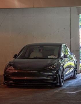 A sleek, dark-colored Tesla car is parked in a dimly lit concrete garage. The electric vehicle is positioned in front of a plain, textured wall, with some natural light coming in from a side opening.