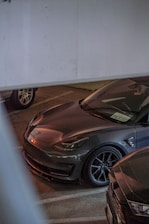 A grey Tesla car is parked in a dimly lit indoor garage. The car has a sleek and modern design, with distinctive headlights and a glossy finish. It is positioned next to another vehicle, with part of a license plate visible through the windshield.