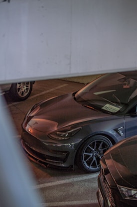 A grey Tesla car is parked in a dimly lit indoor garage. The car has a sleek and modern design, with distinctive headlights and a glossy finish. It is positioned next to another vehicle, with part of a license plate visible through the windshield.