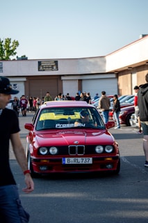 A red BMW car is parked in a crowded area with several people walking around. The car has a license plate that reads 'B RL 737' and the word 'Prosperity' is visible on the windshield. The setting appears to be a car meet or show, with a building in the background and additional cars parked nearby.