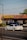A white sports car is driving on a street in front of a Kelly-Moore Paints store. The store has a brown tiled roof and a sign with yellow text on a blue background. Other vehicles, including a black SUV, are parked near the store. There are trees on the right side of the image and a streetlight pole visible on the left side.