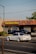 A white sports car is driving on a street in front of a Kelly-Moore Paints store. The store has a brown tiled roof and a sign with yellow text on a blue background. Other vehicles, including a black SUV, are parked near the store. There are trees on the right side of the image and a streetlight pole visible on the left side.