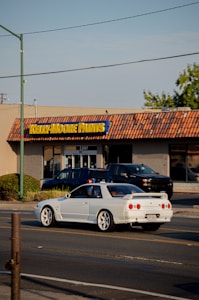 A white sports car is driving on a street in front of a Kelly-Moore Paints store. The store has a brown tiled roof and a sign with yellow text on a blue background. Other vehicles, including a black SUV, are parked near the store. There are trees on the right side of the image and a streetlight pole visible on the left side.